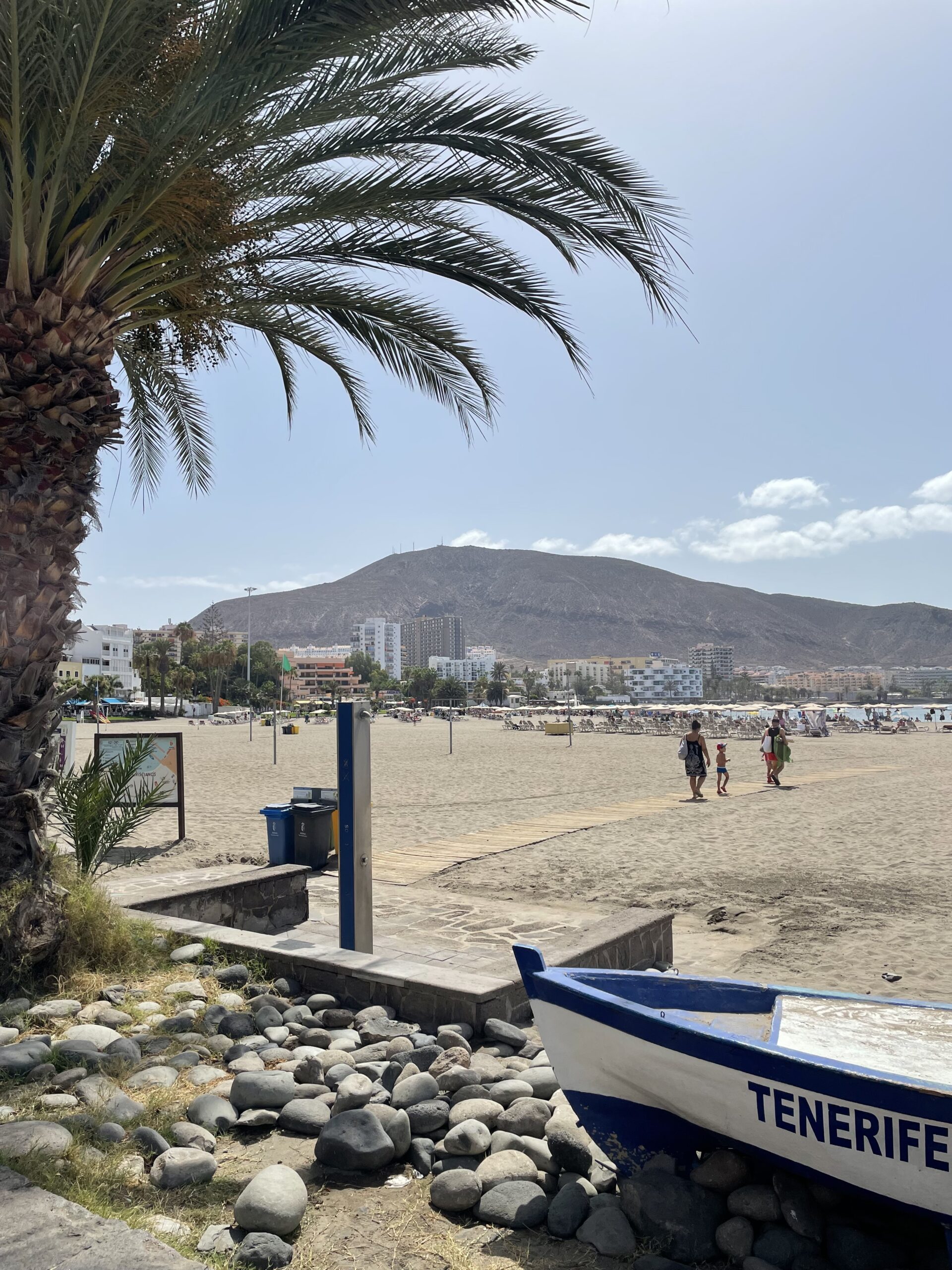 Los Cristianos wide beach a sunny day with blue sky with a mountain in the background. In the foreground a fishing boat named Teneriffa and a palm.